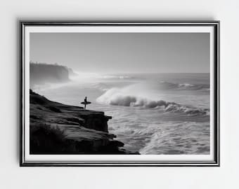 May include: Black and white framed print of a surfer standing on a cliff, holding a surfboard, overlooking the ocean. The waves are crashing, and the sky is overcast. The image is a landscape photograph.