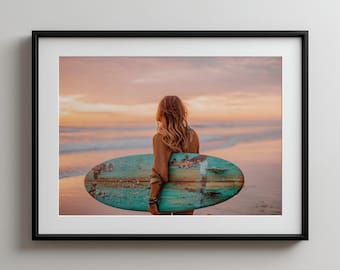 May include: A framed print featuring a person standing on a beach at sunset, holding a weathered, turquoise surfboard. The person is facing the ocean, with the sun setting in the background, casting a warm glow.