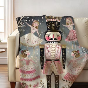 May include: A decorative throw blanket featuring a whimsical illustration of a nutcracker and ballerinas. The blanket has a light pink, black, and white color scheme. The design includes a large nutcracker figure and several dancing ballerinas.