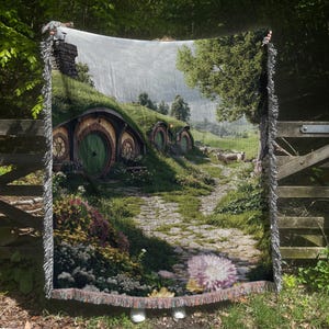 May include: A woven tapestry throw blanket featuring a whimsical scene of hobbit holes nestled in a green hillside with a stone path leading to a field of sheep. The blanket has a fringe border.