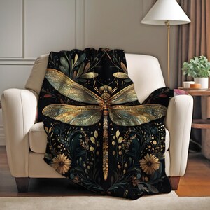 May include: A black throw blanket draped over a white armchair. The blanket features a large, detailed dragonfly design in gold and teal, surrounded by floral and botanical elements. The background is a dark, rich color.
