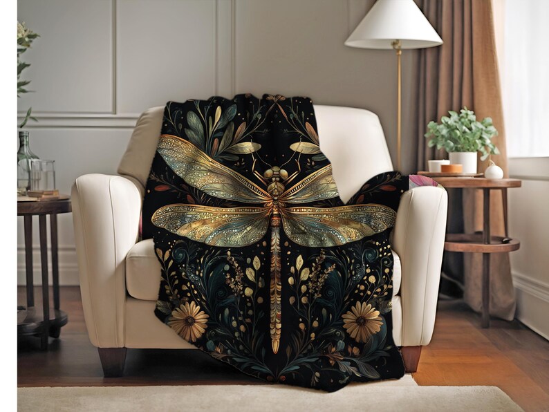 May include: A black throw blanket draped over a white armchair. The blanket features a large, detailed dragonfly design in gold and teal, surrounded by floral and botanical elements. The background is a dark, rich color.