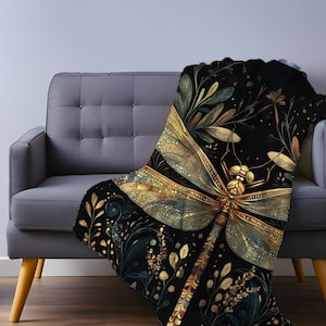 May include: A black throw blanket with a large, detailed dragonfly design in gold and green. The dragonfly is the focal point, surrounded by botanical elements. The blanket is draped over a gray sofa, creating a cozy and decorative accent.