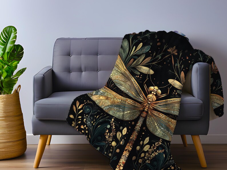 May include: A black throw blanket with a large, detailed dragonfly design in gold and green. The dragonfly is the focal point, surrounded by botanical elements. The blanket is draped over a gray sofa, creating a cozy and decorative accent.