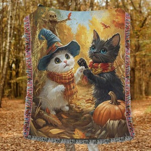 May include: Woven throw blanket featuring a whimsical autumn scene with two kittens. One kitten wears a witch hat and the other a scarf. A pumpkin and a scarecrow are also depicted in the fall foliage. The blanket has fringed edges.