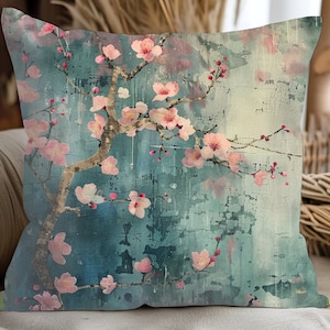 May include: A decorative cushion with a blue and pink floral design. The cushion features a watercolour-style painting of a cherry blossom branch with pink flowers and green leaves. The background is a light blue with a distressed, textured look.