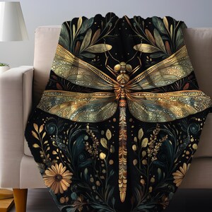 May include: A black throw blanket with a large, detailed dragonfly design in gold and teal. The dragonfly is centered on the blanket, surrounded by floral and leaf patterns in gold and teal. The blanket is draped over a beige sofa.