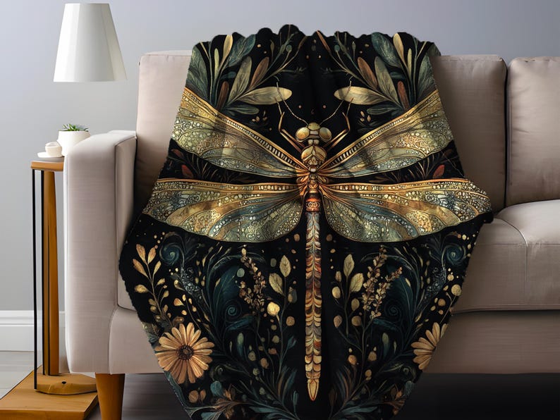 May include: A black throw blanket with a large, detailed dragonfly design in gold and teal. The dragonfly is centered on the blanket, surrounded by floral and leaf patterns in gold and teal. The blanket is draped over a beige sofa.