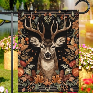 May include: A black garden flag with a detailed illustration of a deer with antlers surrounded by autumn leaves, acorns, and pine cones. The deer has a brown and white coat and is looking directly at the viewer.
