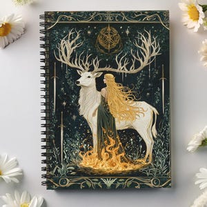 May include: A spiral bound notebook with a dark green cover featuring a white stag with large antlers and a woman with long blonde hair standing beside it. The stag and woman are surrounded by a golden fire and a dark green forest. The cover also features a gold star pattern and a gold circle with a symbol in the center.