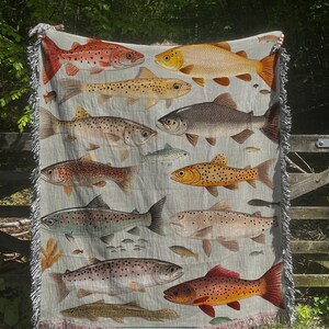 May include: A woven tapestry throw blanket featuring a colorful pattern of various types of fish, including trout, salmon, and bass. The blanket has a light gray background with a fringe border.