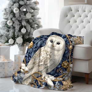 May include: A white owl with gold accents sits on a blue and gold throw blanket with a wintery forest scene. The blanket is draped over a white armchair.