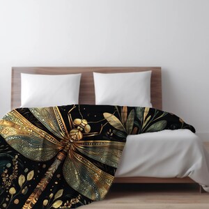May include: A black blanket with a large, detailed dragonfly design in gold and green. The dragonfly is the focal point, with intricate wing patterns. The blanket is draped over a bed with white bedding, creating a striking contrast.