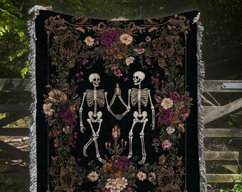 May include: Black woven throw blanket featuring a design of two skeletons holding hands, surrounded by floral arrangements in shades of purple, cream, and brown. The blanket has a fringed edge and is displayed outdoors.