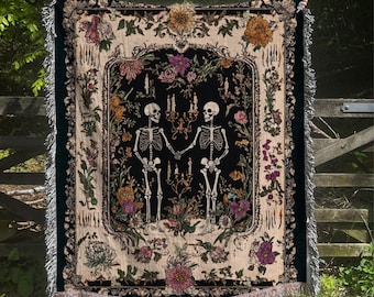 May include: A woven tapestry blanket featuring a black central panel with two skeletons holding hands, surrounded by floral and candle designs. The blanket has a cream-colored border with fringe and is decorated with various flowers in shades of purple, orange, and yellow.