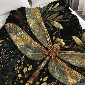 May include: A decorative blanket featuring a detailed illustration of a dragonfly with intricate wing patterns in shades of gold, teal, and brown. The dragonfly is set against a dark background with floral and leaf accents, creating a nature-inspired design.