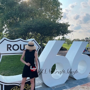 May include: A person wearing a black dress and sandals, holding a red and white crocheted purse. The background features a Route 66 sign and large white numbers 66. The scene is outdoors on a sunny day.