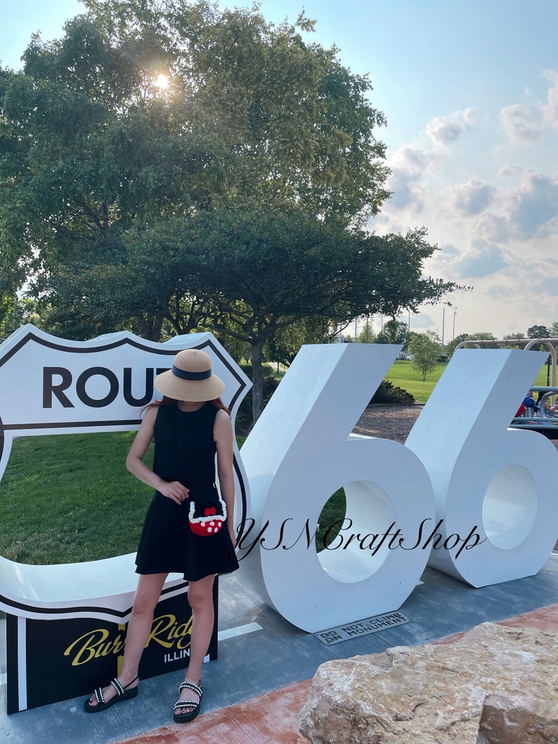 May include: A person wearing a black dress and sandals, holding a red and white crocheted purse. The background features a Route 66 sign and large white numbers 66. The scene is outdoors on a sunny day.