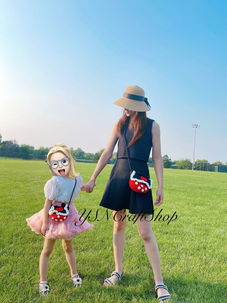 May include: Two people holding hands, each wearing a red, black, and white crochet purse. The purses have a round shape with a black strap. The text "YSN Craft Shop" is visible. The people are standing on a green grassy field.