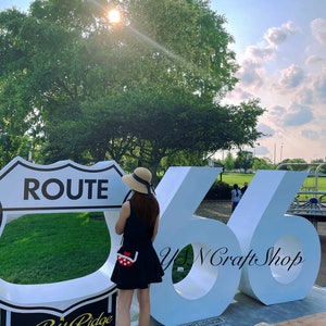 May include: A person stands near a Route 66 sign and large white numbers. The person wears a black dress, sandals, and a straw hat with a blue ribbon. A small red and white polka dot purse is visible. The sign reads "Burr Ridge, Illinois."