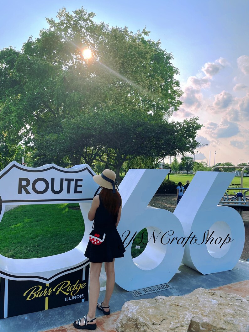 May include: A person stands near a Route 66 sign and large white numbers. The person wears a black dress, sandals, and a straw hat with a blue ribbon. A small red and white polka dot purse is visible. The sign reads "Burr Ridge, Illinois."
