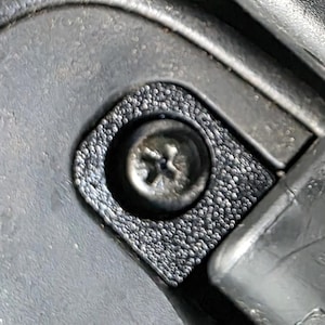 May include: Close-up of a black plastic component with a textured surface and a silver screw. The screw is centered within a textured, rounded square. The surrounding plastic has a smooth, glossy finish.
