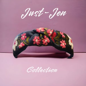 May include: A dark blue headband with a knotted design and embroidered pink and white flowers. The headband says "Just-Jen Collection" in white text.