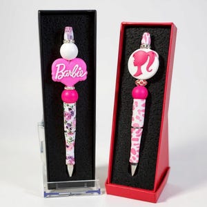 May include: Two decorative pens in display boxes. One pen features a pink heart with the word "Barbie" and a floral design. The other pen has a pink silhouette and a pink and white pattern. Both pens have a silver tip.
