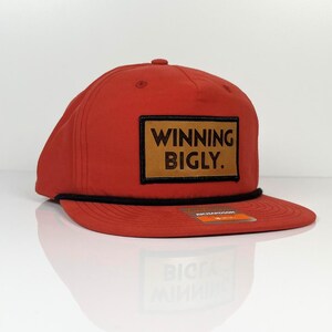 May include: Red baseball cap with a black rope detail and a brown leather patch that says "WINNING BIGLY."
