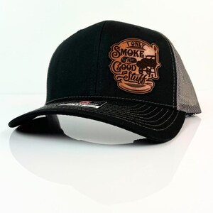 May include: Black trucker hat with a gray mesh back and a curved brim. The hat features a brown leather patch with the text "I Only Smoke The Good Stuff" and a graphic of a smoker.