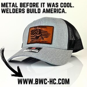 May include: A gray and black trucker hat with a brown leather patch that says "The Original Heavy-Metal Artist". The patch features a silhouette of a welder with an American flag in the background. The hat also has the text "Welders Build America" on the front.