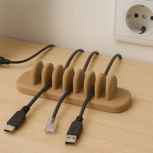 May include: A light brown cable organizer with six rounded slots, holding several black cables. The organizer sits on a light wood surface, with a laptop and a wall outlet visible in the background. The cables include USB and Ethernet connectors.