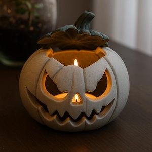 Könnte beinhalten: Ein grauer Keramik-Kürbis-Kerzenhalter mit geschnitztem Gesicht und grünem Blattdeckel. Zwei brennende Kerzen im Inneren werfen einen warmen Schein durch Augen und Mund, ideal für Halloween-Dekorationen.