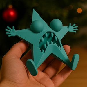 May include: A teal-colored star-shaped figurine with a cartoonish design. It features large, round eyes, a wide open mouth with teeth, and outstretched arms and legs. The figurine is held in a hand, with a blurred background.