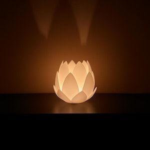 Op de afbeelding: Een witte lamp in de vorm van een lotusbloem gloeit tegen een donkere achtergrond. De bloemblaadjes van de lamp worden van binnenuit verlicht, waardoor zacht licht en schaduwen ontstaan. Het algehele effect is een warme, uitnodigende sfeer.