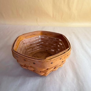 May include: A hexagonal woven wooden basket featuring a light tan natural finish with visible wood grain and metal fasteners along the rim. The interior contains a clear, molded plastic liner that mirrors the hexagonal shape of the basket structure.