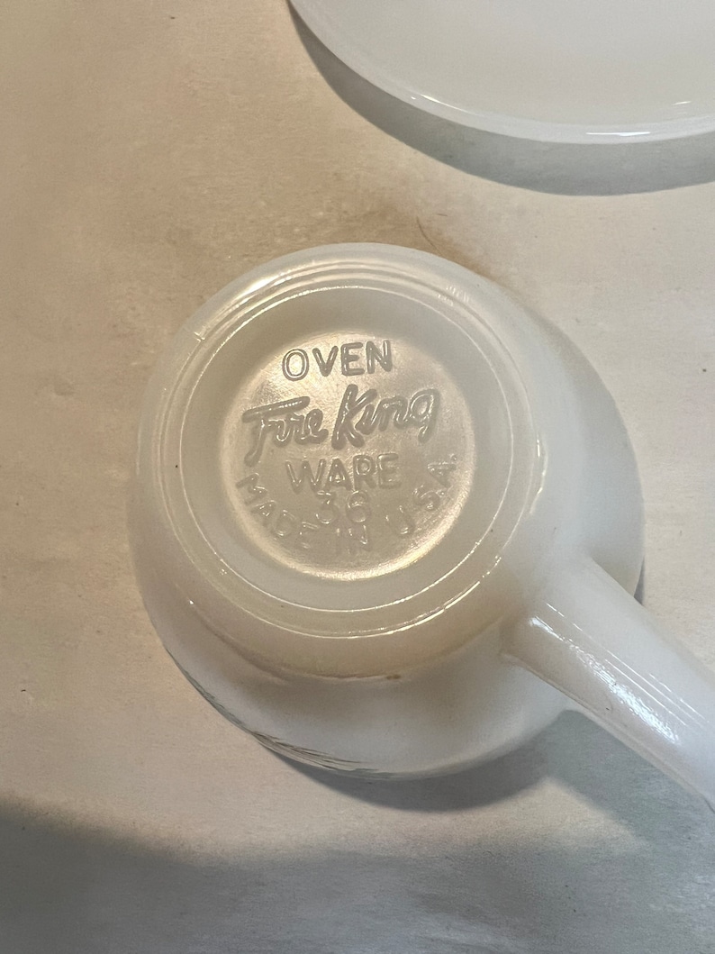 May include: White ceramic mug with a handle. The bottom of the mug has the words "Oven Fire King Ware" printed on it.