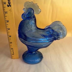 May include: A cobalt blue glass rooster-shaped covered dish with a textured feather pattern and a clear glass comb. A yellow wooden ruler is positioned vertically next to the object, showing a total height of approximately 8.5 inches.