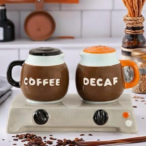 May include: A ceramic coffee and decaf salt and pepper shaker set. The brown shakers are shaped like coffee mugs with black and orange handles. The word "COFFEE" is on one shaker and "DECAF" on the other. They sit on a light gray base.