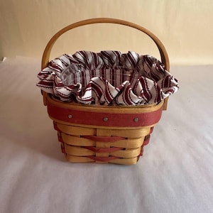 May include: A small, woven basket with a wooden handle and a red rim. The basket is lined with a ruffled fabric featuring red and white stripes. The basket is designed for decorative or storage purposes.