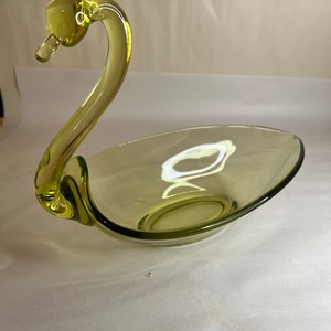 May include: A green glass swan shaped dish. The dish is oval and has a curved lip. The swan's neck is curved and the head is tilted to the side.
