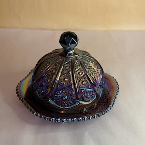 Op de afbeelding: Iridescent glazen botervloot met een gewelfde deksel en een decoratieve basis. De schaal heeft ingewikkelde reliëfpatronen en een facetgeslepen zwarte knop. De kleuren verschuiven van paars naar blauw en groen.