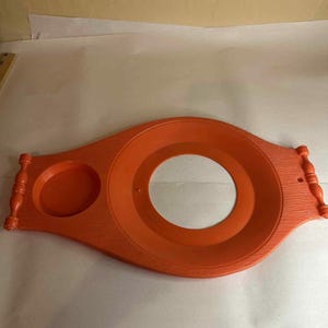 May include: An orange wooden serving tray with a large central circular opening and a smaller circular indentation. The tray has decorative handles and a textured surface.