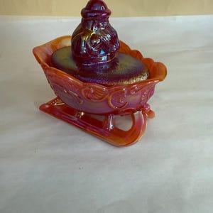 Puede incluir: Un cuenco de cristal iridiscente rojo y naranja con forma de trineo. La tapa presenta una figura de Papá Noel. El cuenco tiene volutas decorativas y está diseñado para uso festivo.