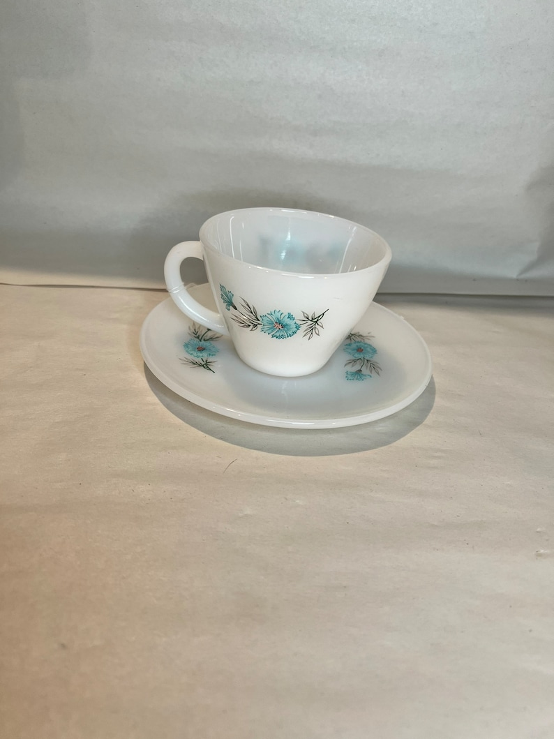 May include: A white teacup and saucer set with a blue floral design. The cup has a handle and the saucer has a scalloped edge.