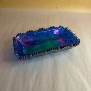 Puede incluir: Un plato rectangular de vidrio con un acabado iridiscente en azul y morado, que presenta un patrón geométrico facetado en los lados y un borde festoneado y dentado. La superficie refleja la luz con tonos metálicos cambiantes.
