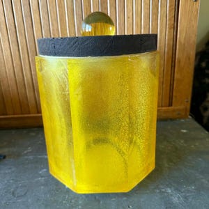 May include: A yellow, hexagonal jar with a black rim and a clear, yellow ball-shaped knob. The jar has a textured appearance and is made of a translucent material. The background features a wooden slatted surface.