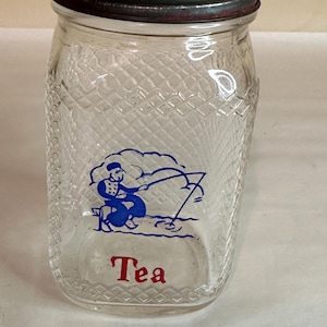 May include: A clear glass jar with a metal lid and a blue and white illustration of a person fishing. The jar has a diamond pattern and the word "Tea" in red lettering.