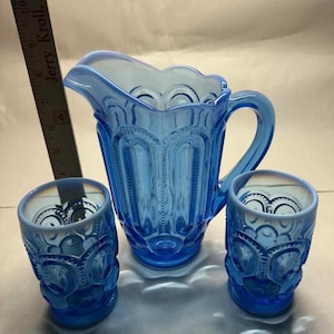Op de afbeelding: Een vintage blauwe glazen kan en twee bijpassende glazen. De kan heeft een handvat en een geschulpte rand. De glazen en de kan hebben een decoratief patroon. De set is gemaakt van blauw glas.