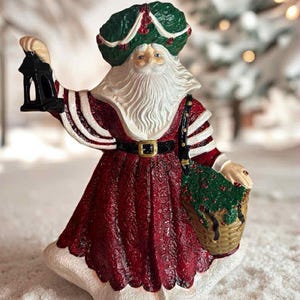 May include: A Christmas-themed Santa Claus figurine, wearing a red and white robe with a black belt. He holds a black lantern and a basket of greenery. The figure has a white beard and a green turban-style hat. Festive decoration.